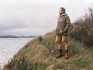 Man standing on a grassy hill by a body of water wearing a green jacket and brown shorts.